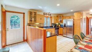 A kitchen with wooden cabinets, a stainless steel refrigerator, and a dining area with a table and chairs. - Upper Valley Retreat on 15 Acres Near Dartmouth (Meriden)
