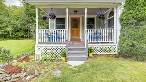 A two-story house with a porch, a front door, and a small garden area with a stone border and potted plants. - Upper Valley Retreat on 15 Acres Near Dartmouth (Meriden)