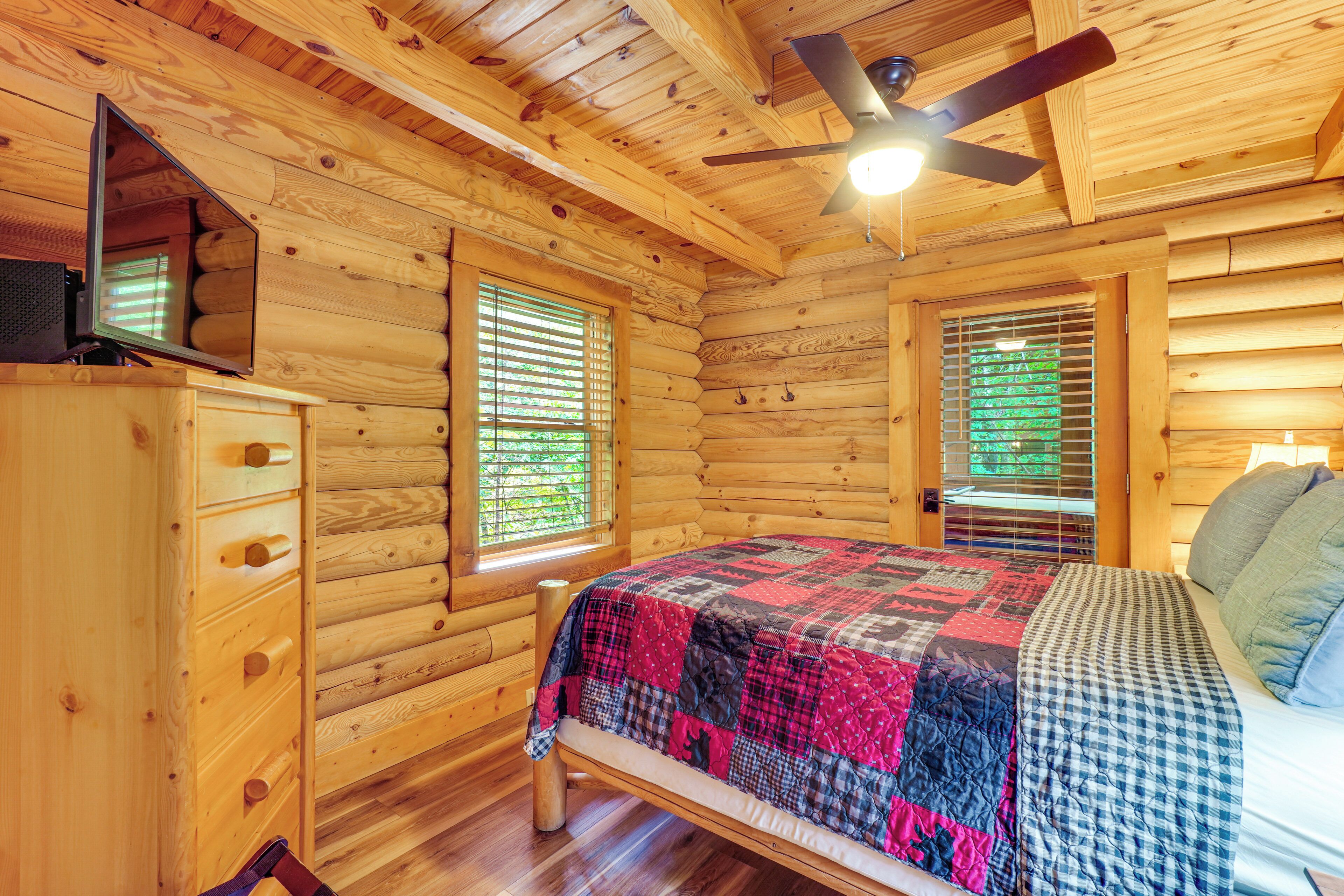 Ein Schlafzimmer in einer Blockhütte mit einem Bett, einer Kommode, einem Deckenventilator und zwei Fenstern mit Jalousien.