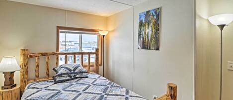 A bedroom with a wooden bed frame, a quilt, a window with a view, a painting on the wall, and a lamp.