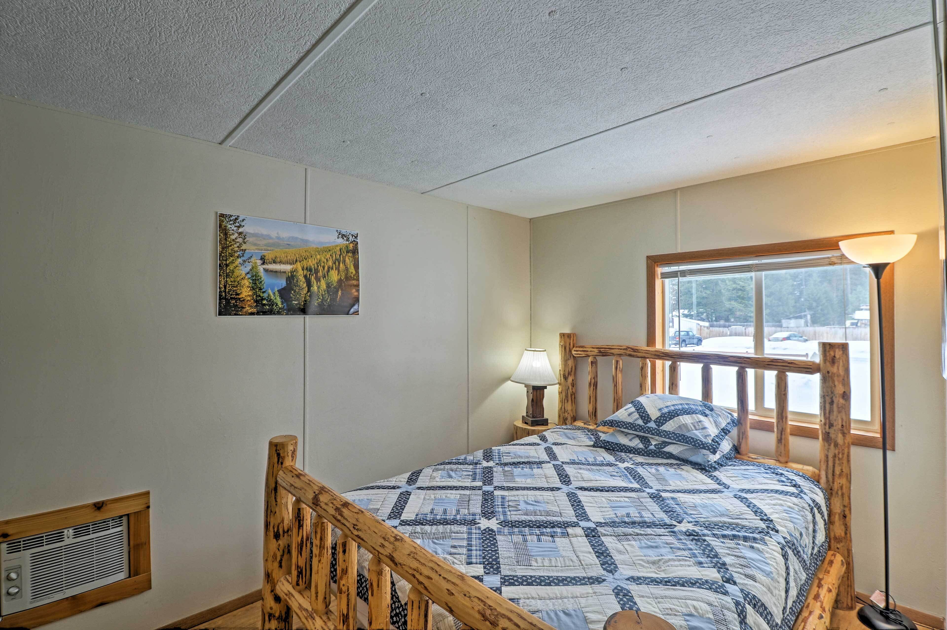 A bedroom with a wooden bed frame, a quilted bedspread, a nightstand with a lamp, a window with a view of a snowy landscape, and a wall-mounted heater.