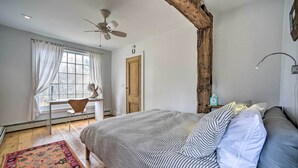 A bedroom with a large bed, a desk, and a chair. There is a window with curtains, a ceiling fan, and a wooden beam. - Charming Cottage w/ Yard - 2 Mi to Tinker St! (West Hurley)