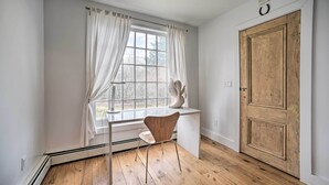 A room with a wooden floor, a white desk with a chair, a window with white curtains, and a wooden door. - Charming Cottage w/ Yard - 2 Mi to Tinker St! (West Hurley)
