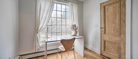 A room with a wooden floor, a white desk with a chair, a window with white curtains, and a wooden door.