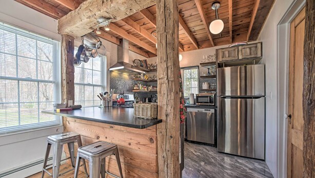 A rustic kitchen with wooden beams, a large island with stools, stainless steel appliances, and a view through large windows. - Charming Cottage w/ Yard - 2 Mi to Tinker St! (West Hurley)