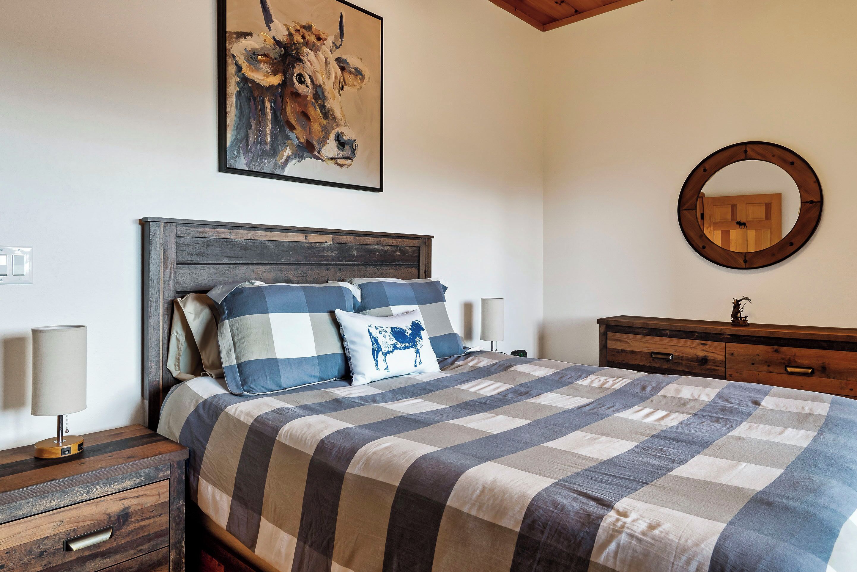 Une chambre à coucher avec un lit, une table de chevet en bois, une commode, un miroir rond et un tableau représentant une vache accroché au mur.