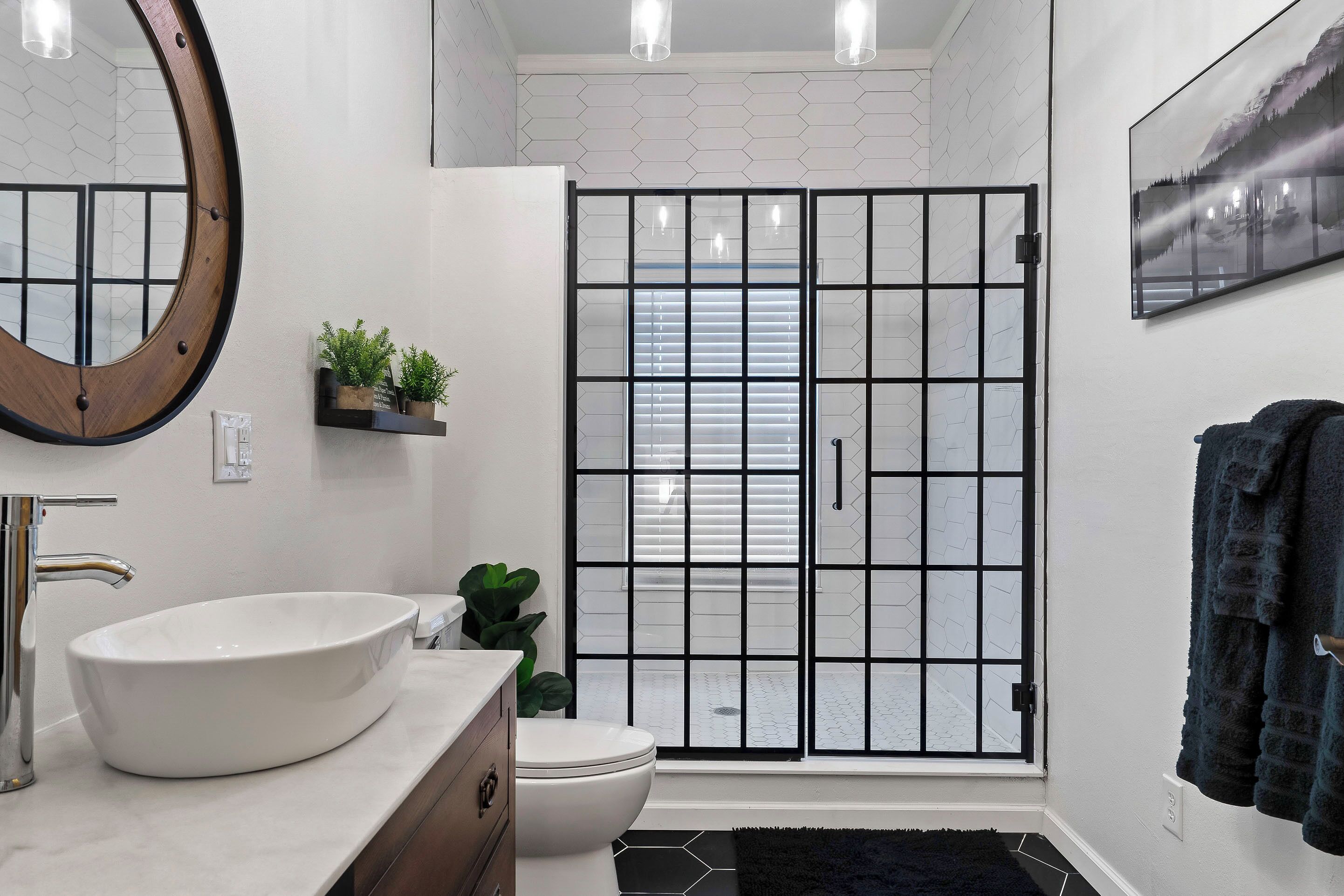 Un bagno con un lavandino bianco, uno specchio tondo, un water e una porta in vetro con cornice nera che dà accesso alla zona doccia.