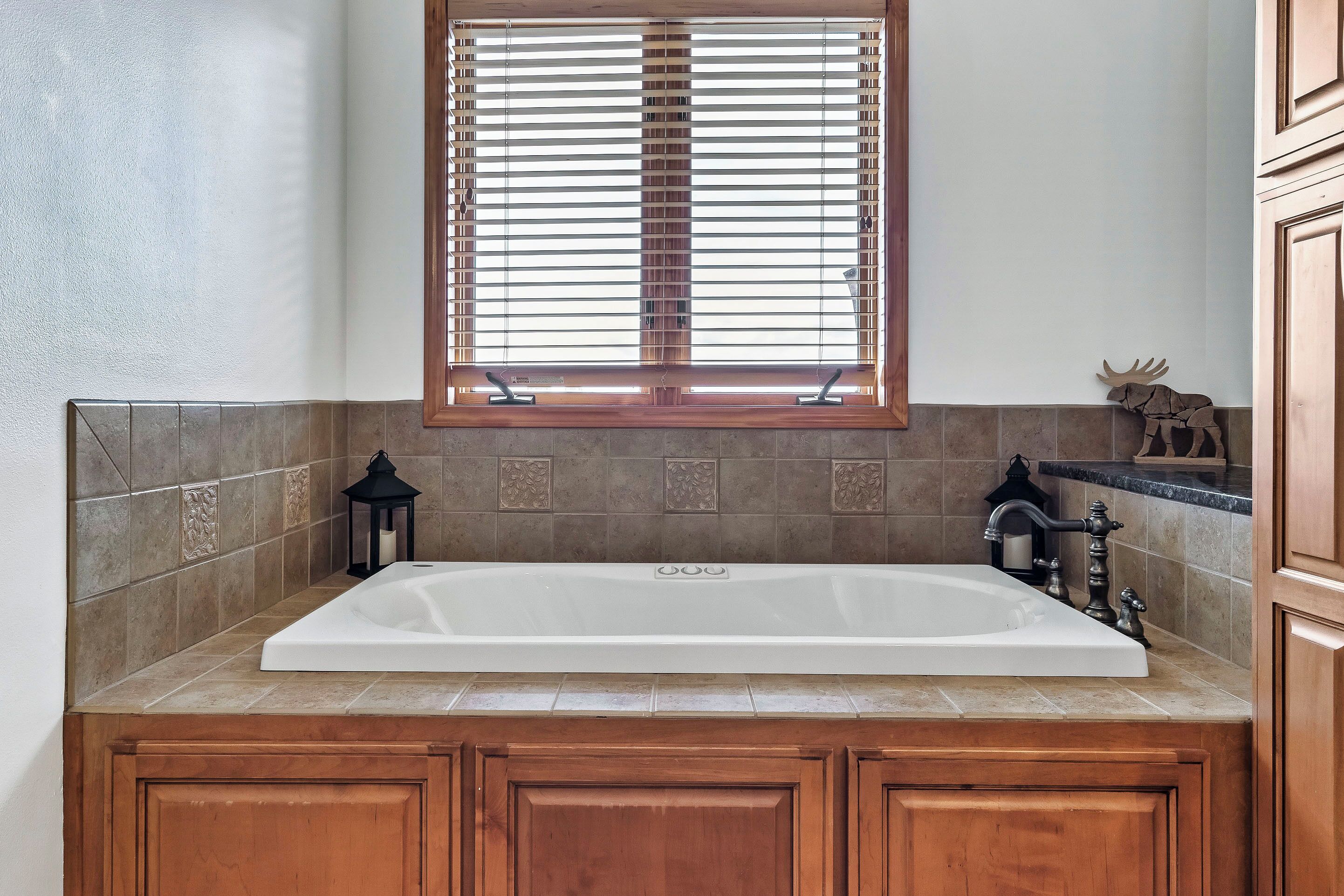 A bathroom with a large white bathtub, wooden cabinets, a window with blinds, and a decorative moose figure.