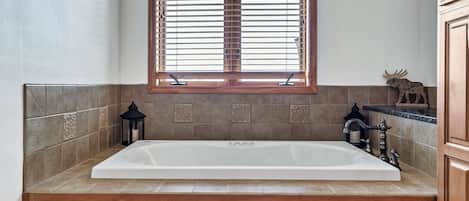 A bathroom with a large white bathtub, wooden cabinets, a window with blinds, and a decorative moose figure.