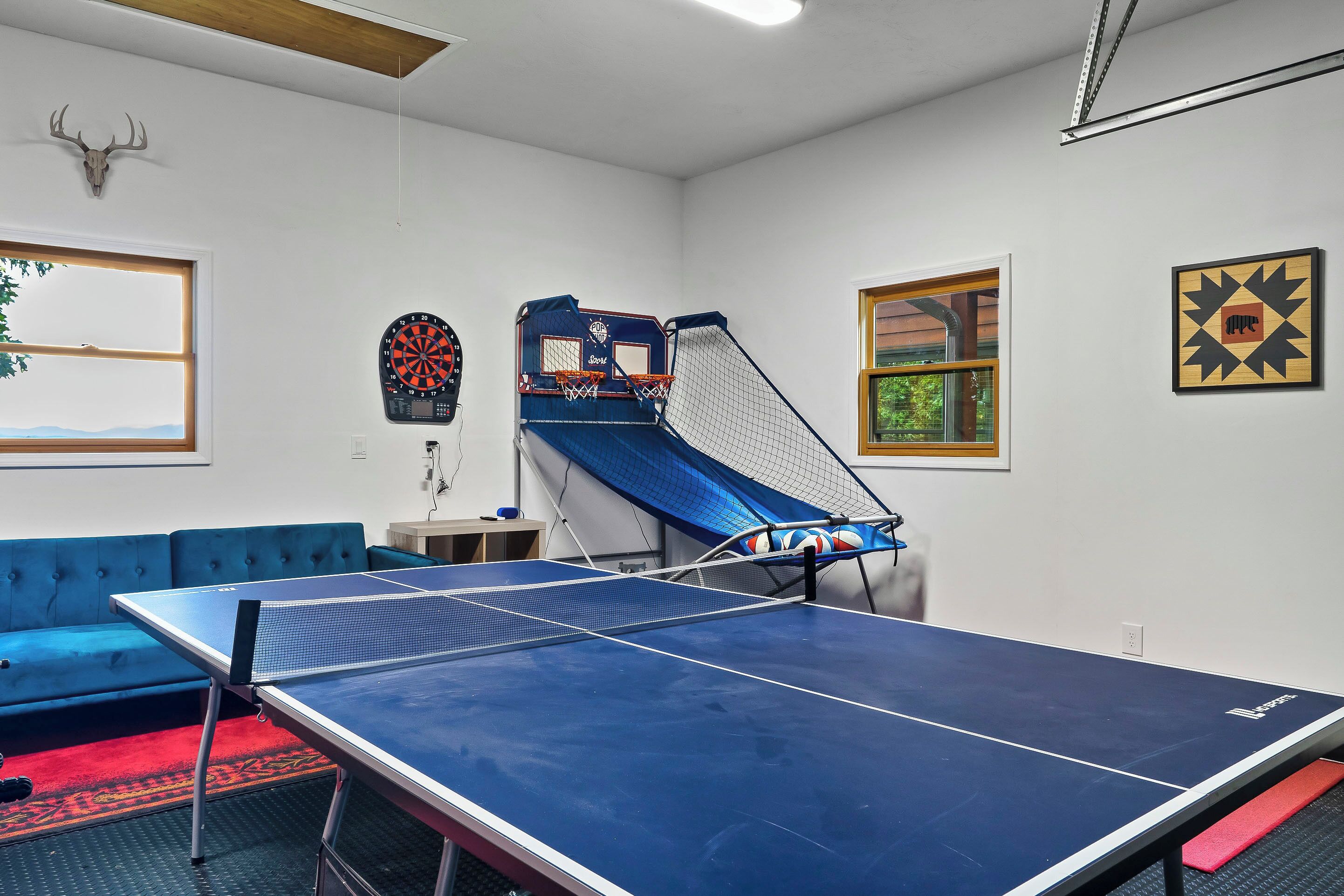 A room with a ping pong table, a blue couch, a dartboard, and a basketball hoop.