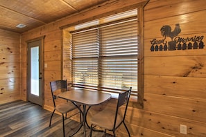 A wooden interior with a dining area featuring a table and chairs, a window with blinds, and a wall with a rooster and 'Good Morning' sign. - Smoky Mtn Log Cabin w/ Hot Tub & Panoramic Views! (Sevierville)