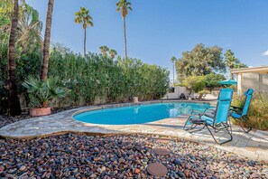 A swimming pool surrounded by pebbles, with palm trees and a patio area with chairs. - Scottsdale Adobe Home w/ Backyard Oasis! (Scottsdale)