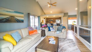 A spacious living room with a high ceiling, a kitchen in the background, and a view of the outside.