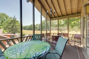 A covered outdoor seating area with a table and chairs, large windows, and a view of the surrounding landscape.