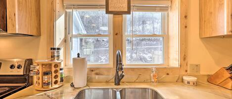 A kitchen with wooden cabinets, a double sink, a window with a view of trees, and a hanging light fixture.