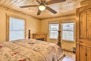 A bedroom with a wooden ceiling, a ceiling fan, a bed with a vintage-themed bedspread, a fireplace, and large windows. - Quaint Appalachian Getaway ~ 2 Mi to Boone! (Boone)