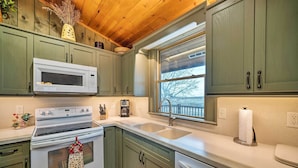 A kitchen with green cabinets, a white microwave, a stove, a sink, and a window with a view of trees. - Secluded Retreat w/ Wood Stove, 11 Mi to Bozeman! (Gallatin Gateway)