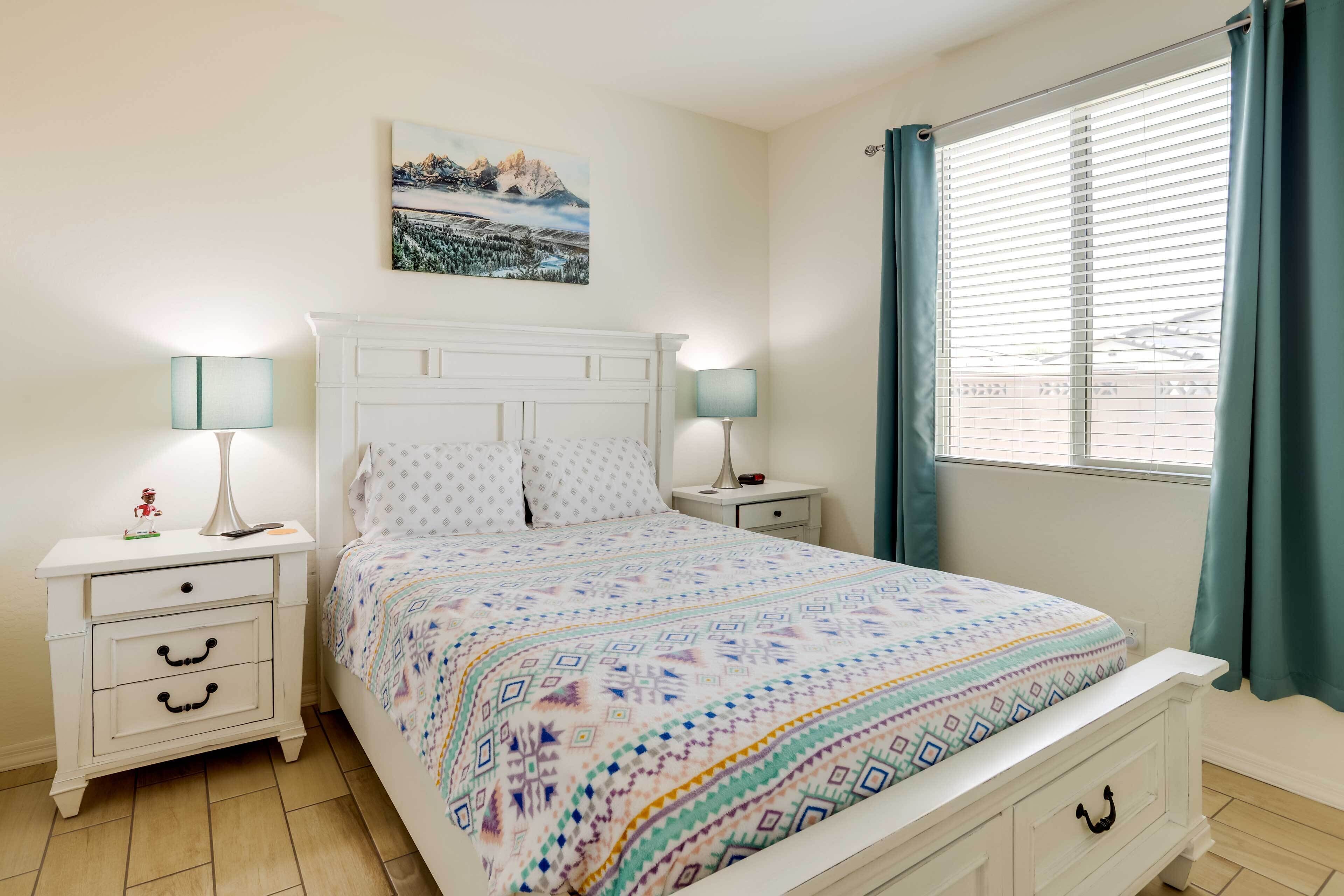 Quarto com cama, duas mesas de cabeceira, um criado-mudo, um quadro na parede e uma janela com cortinas.