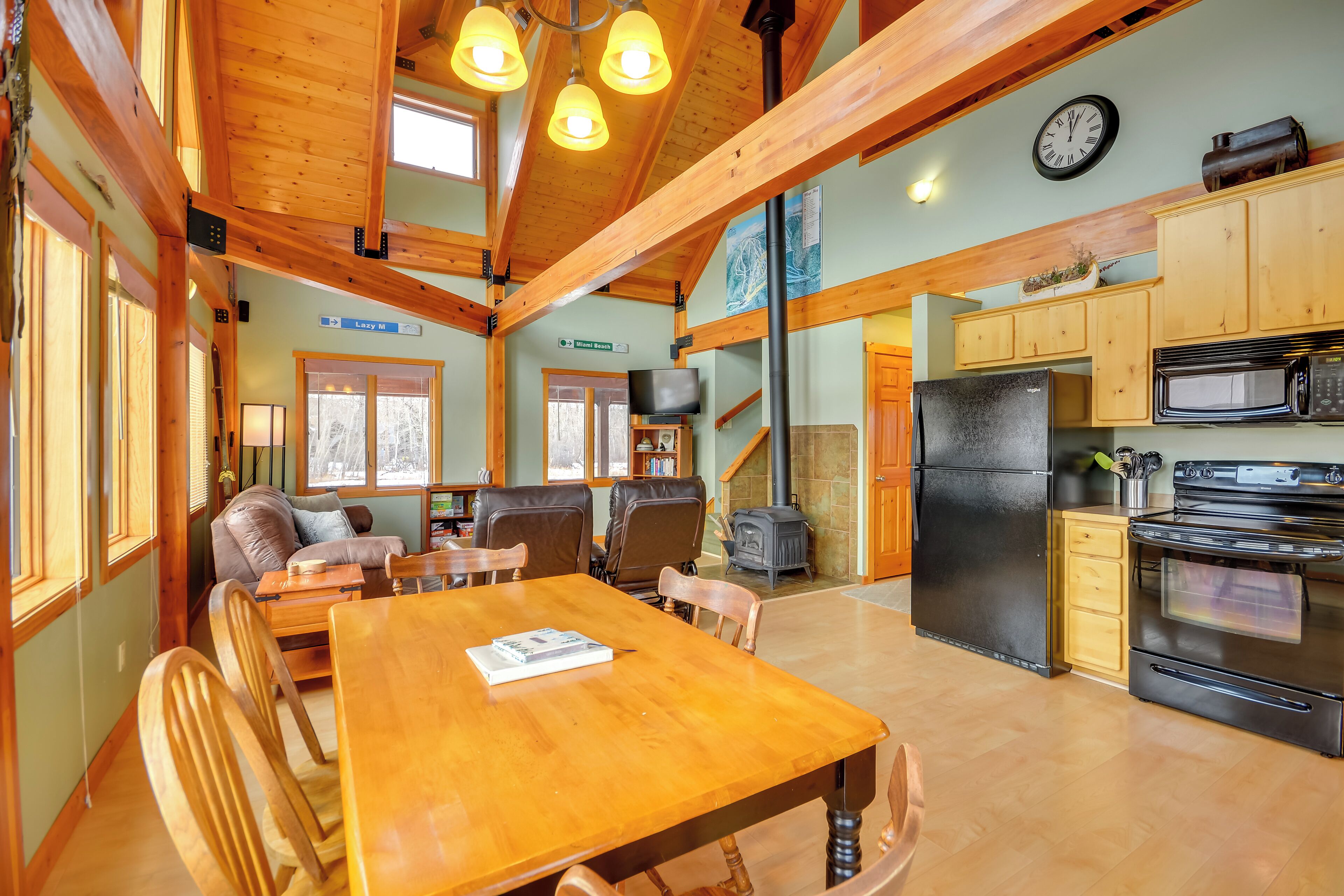 A cozy cabin interior with a wooden dining table, chairs, a fireplace, a kitchen with modern appliances, and a large window.