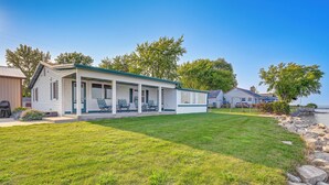 A single-story mobile home with a covered porch, a well-maintained lawn, and a rocky shoreline in the background.