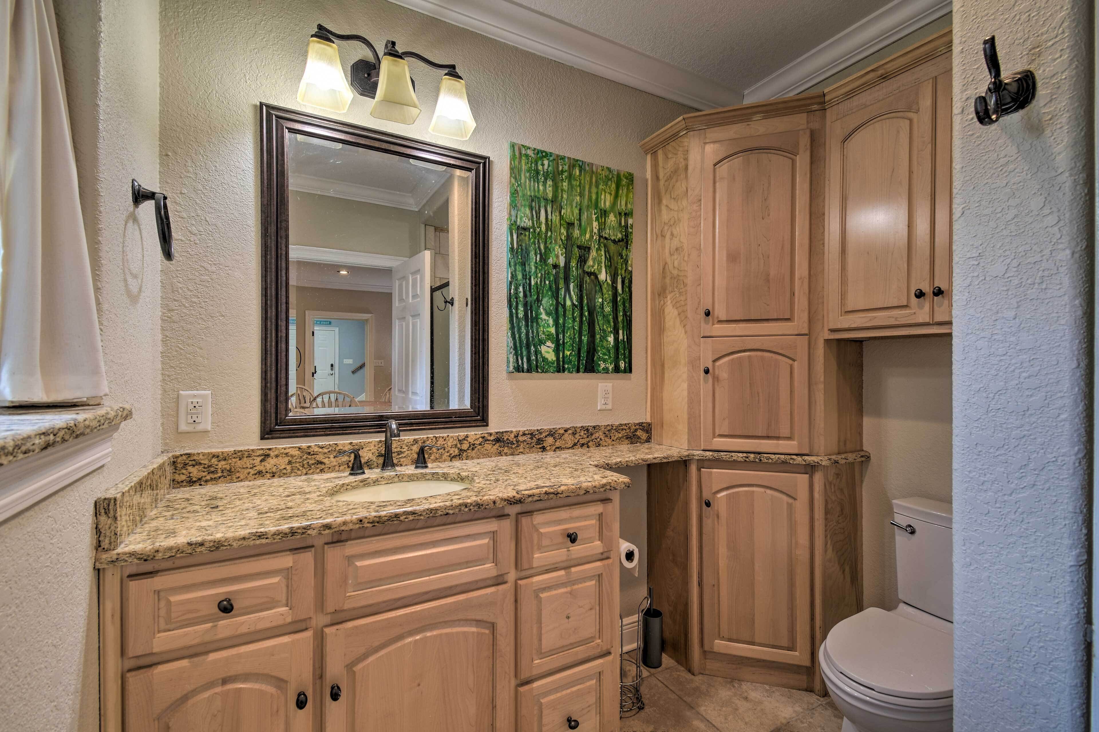 A bathroom with a granite countertop sink, wooden cabinets, a large mirror with three lights, and a green forest-themed wall art.
