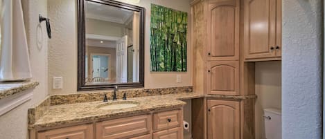 A bathroom with a granite countertop sink, wooden cabinets, a large mirror with three lights, and a green forest-themed wall art.