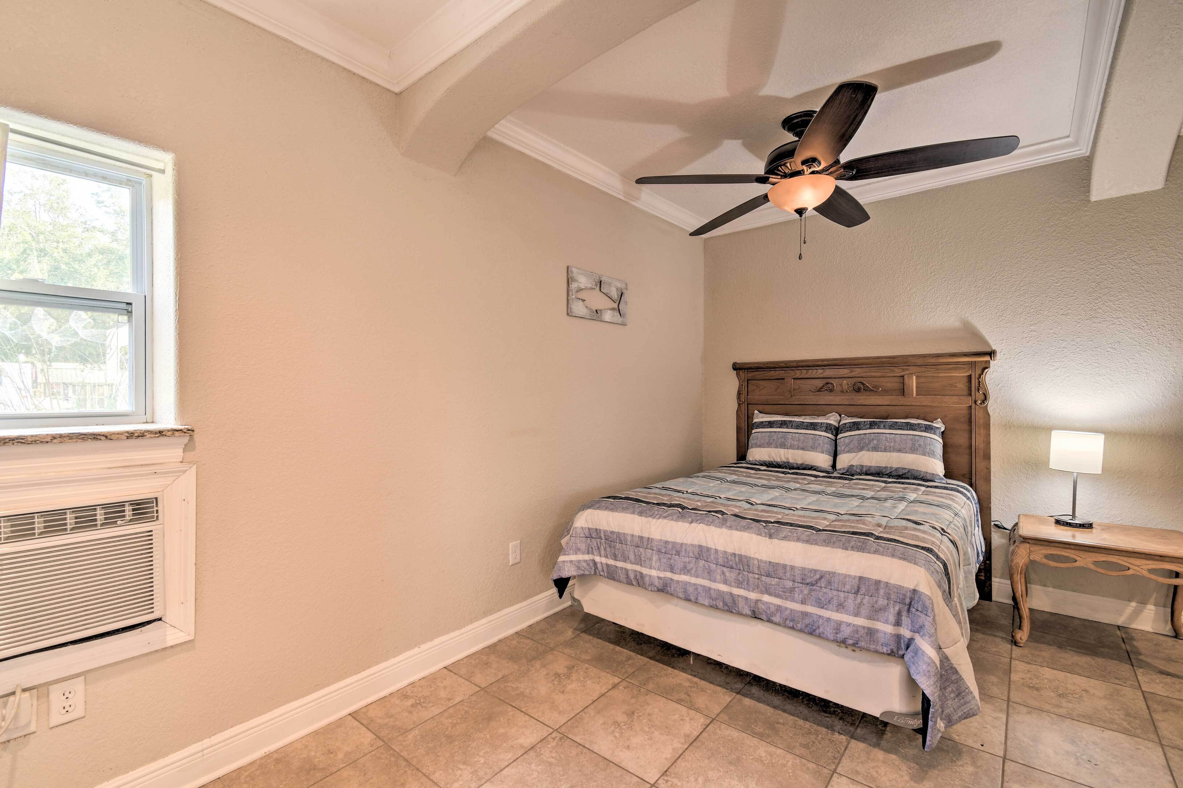 A bedroom with a bed, a ceiling fan, a nightstand, a lamp, a window, and a wall-mounted heater.