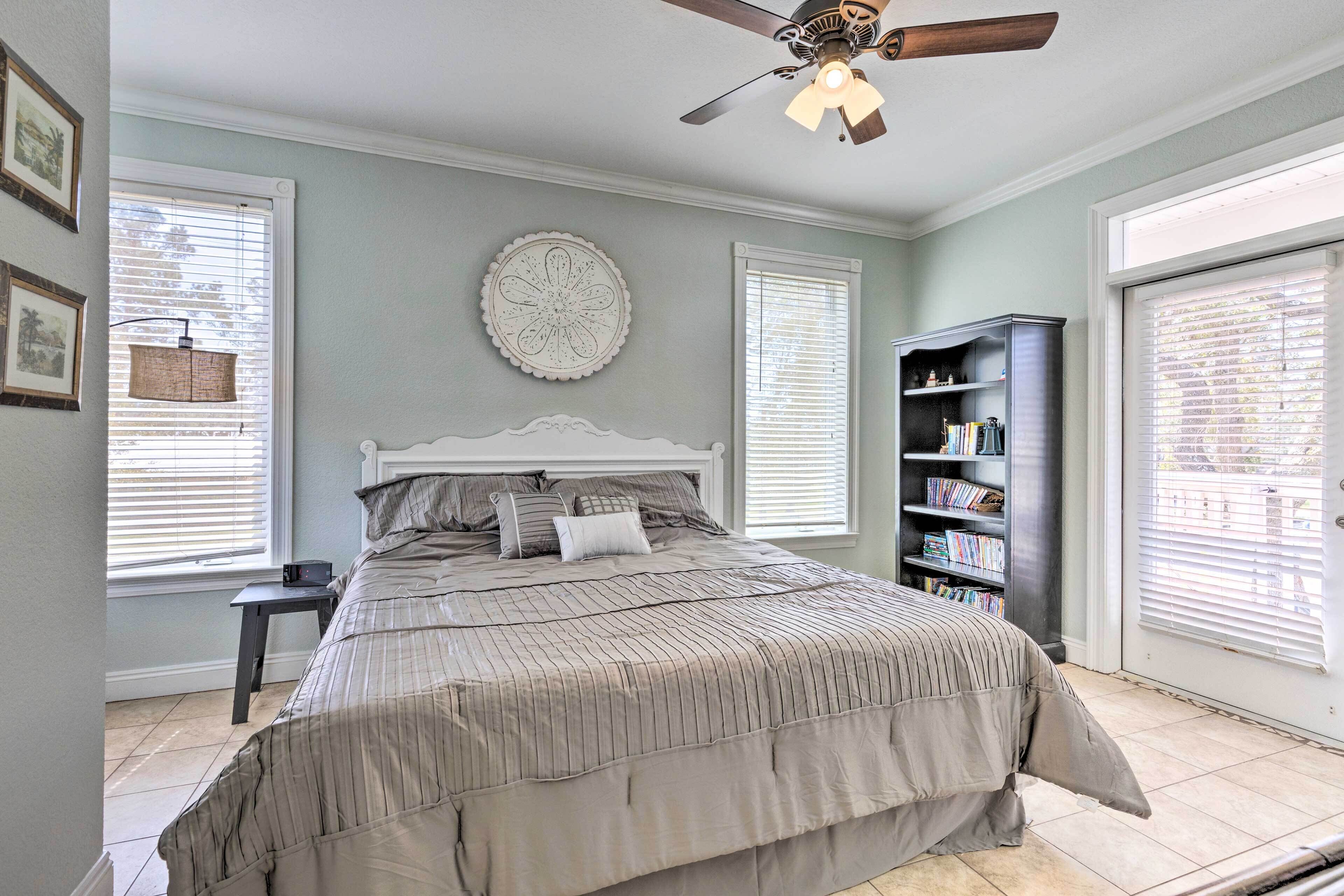 A bedroom with a large bed, a ceiling fan, a bookshelf, and a window with blinds.
