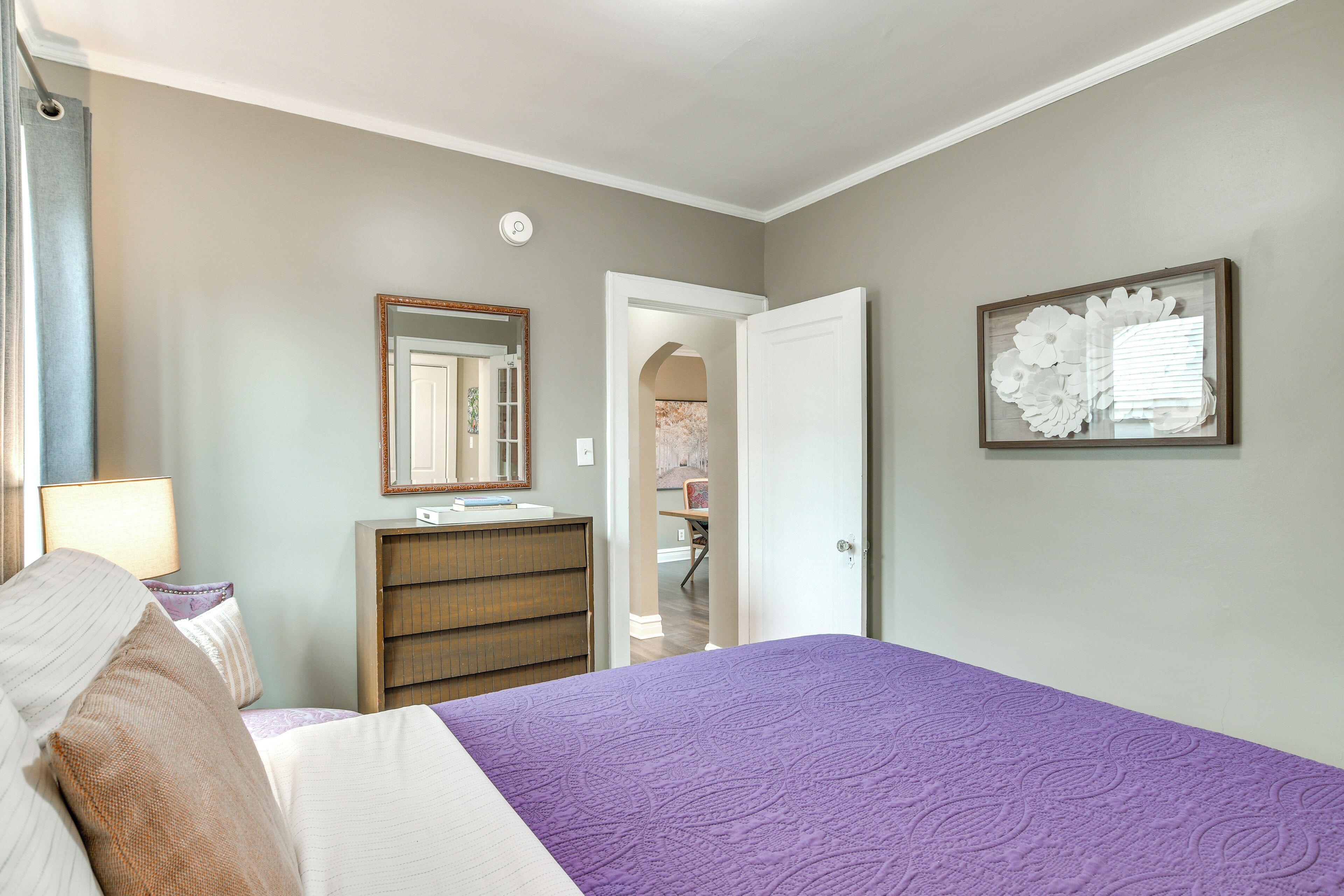 Ein Schlafzimmer mit Bett, Kommode, Spiegel und einer Tür zu einem weiteren Raum.