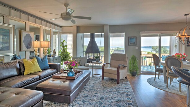 A spacious living room with a leather sofa, a coffee table, and a dining area with chairs. There is a large window offering a view of the outdoors. - Waterfront Lake Travis Home w/ Pool Access! (Leander)