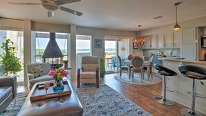 A spacious living room with a sofa, coffee table, and a view of the outdoors. There is a dining area with a table and chairs, and a kitchen with white cabinets and a bar area. - Waterfront Lake Travis Home w/ Pool Access! (Leander)