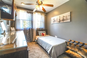 A bedroom with a bed, a desk, a chair, a ceiling fan, and a window with curtains.