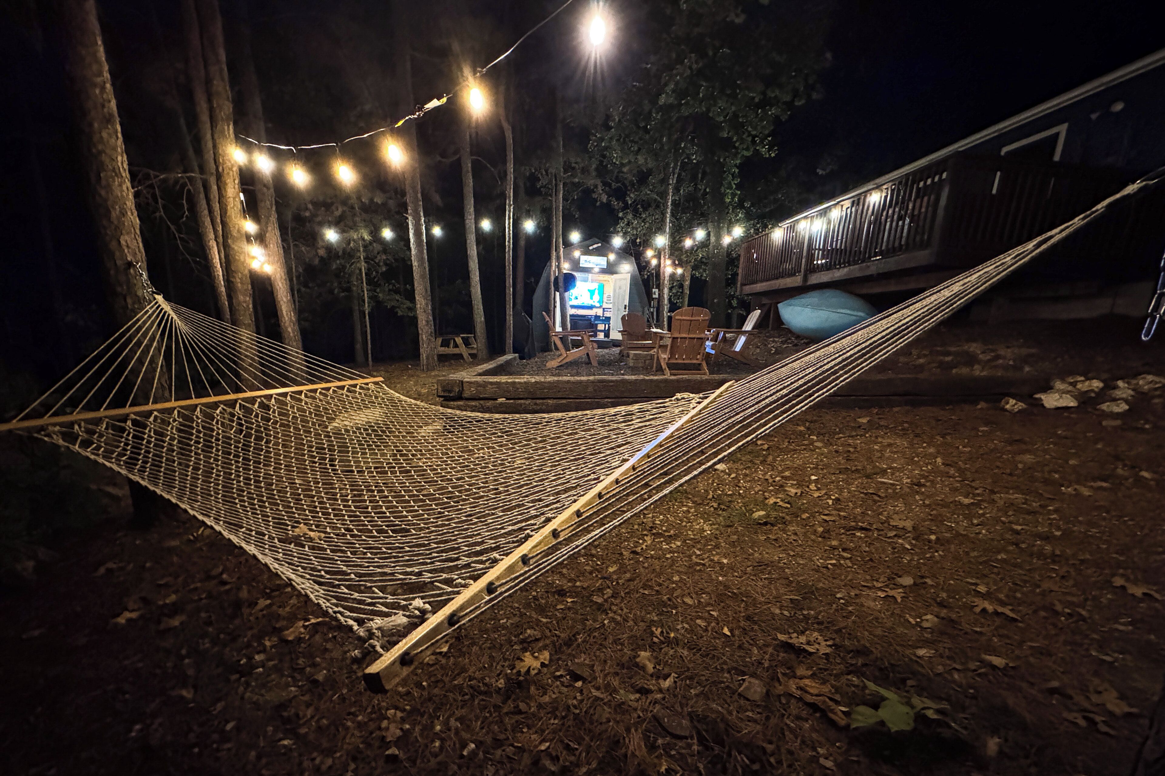 Una hamaca instalada en un área boscosa con luces colgantes, una tienda pequeña y una terraza con sillas y una mesa.