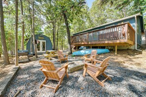 Une cabane dotée d’une terrasse, entourée d’arbres, et aménagée avec des chaises en bois et un foyer.