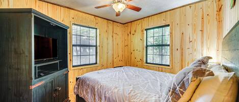 A bedroom with wooden walls, a ceiling fan, a bed with a patterned bedspread, and a TV on a dark wooden cabinet.