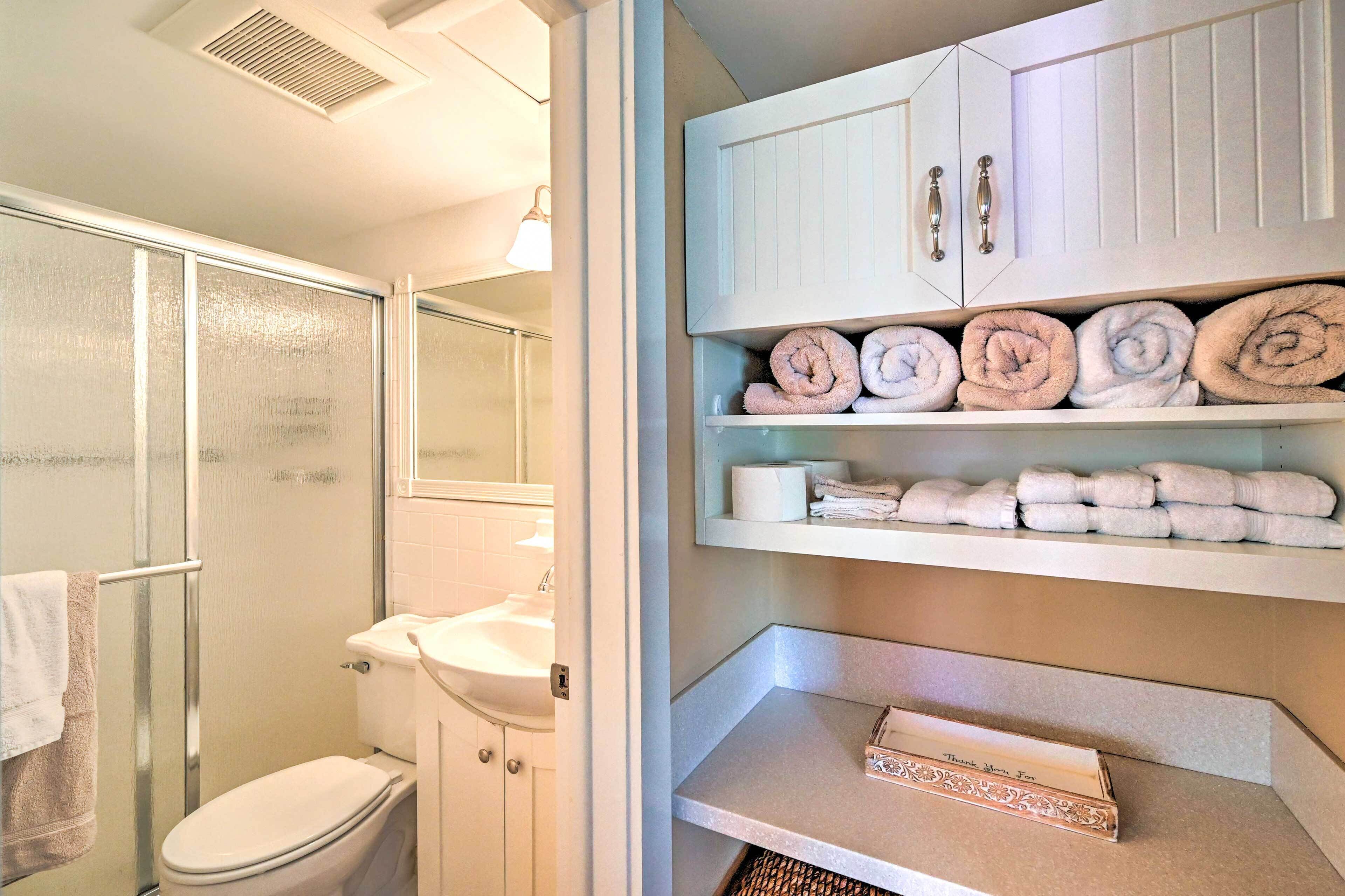 Une salle de bain comprenant des toilettes, un lavabo et un espace douche. On y trouve des étagères avec des serviettes pliées et une boîte contenant un mot de remerciement.