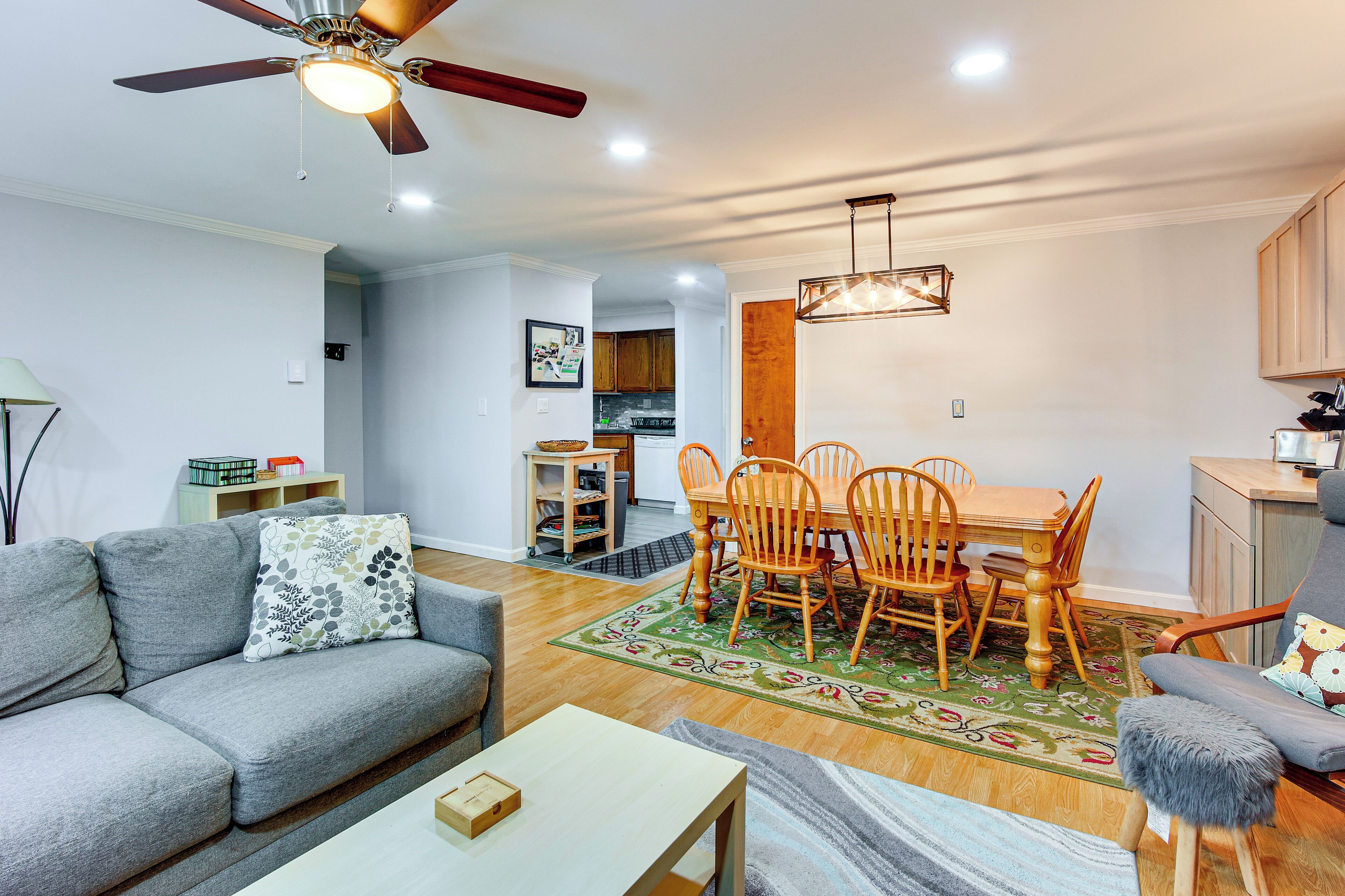 A living room with a sofa, a coffee table, a dining table with chairs, a ceiling fan, and a kitchen area in the background.