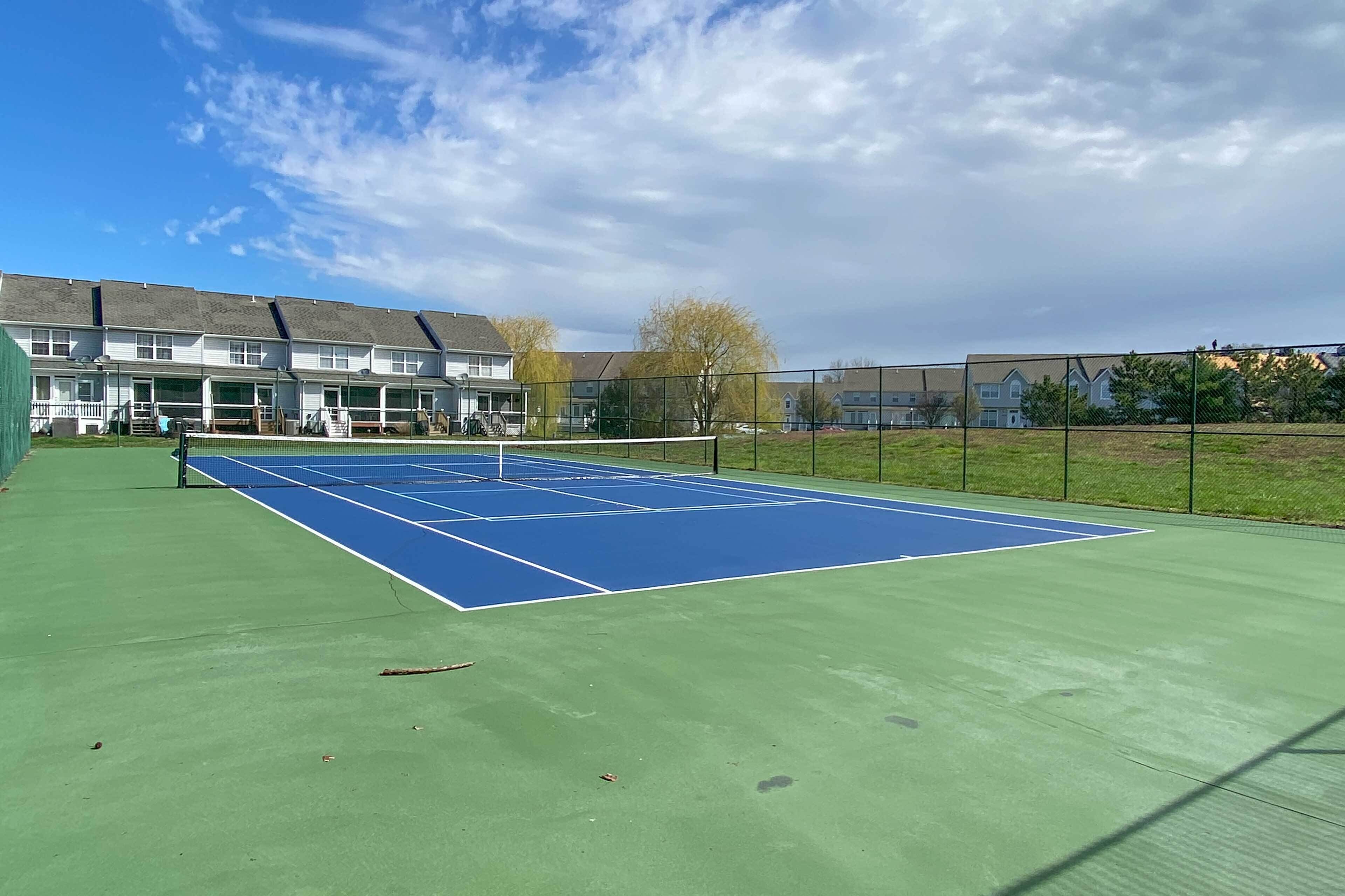 A tennis court with a net, surrounded by a fence, and adjacent to residential buildings.