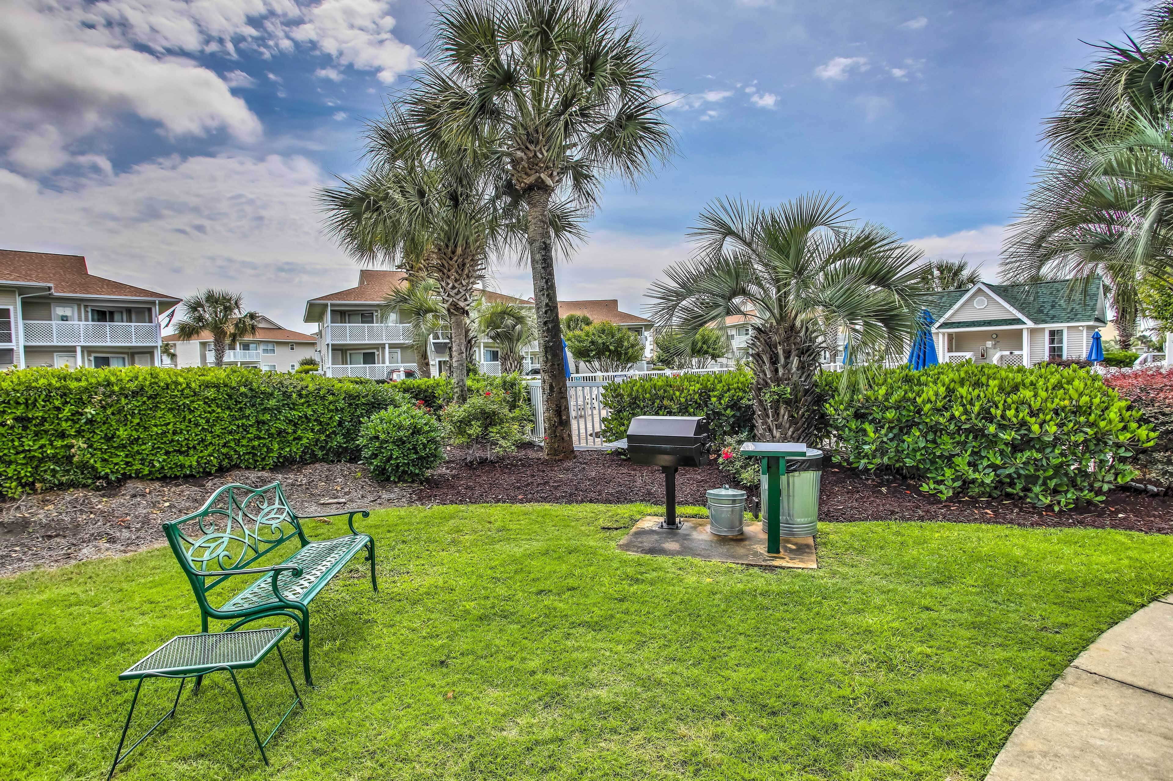 A well-maintained grassy area with a metal bench, a barbecue grill, and a trash can.