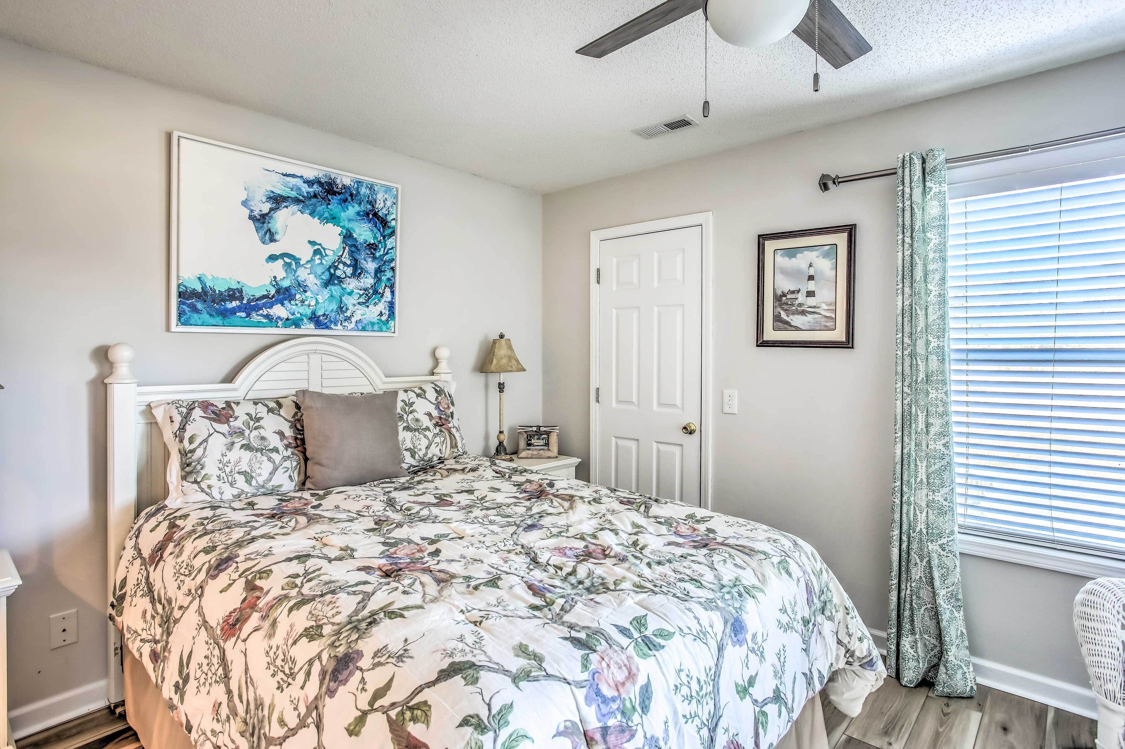 A bedroom with a floral bedspread, a ceiling fan, a framed abstract painting, a door, and a window with blinds.