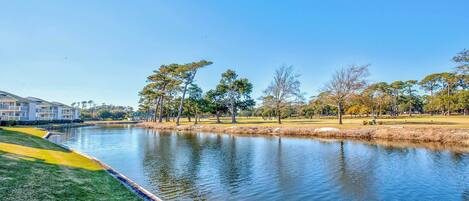Une vue paisible sur un lac, avec un terrain de golf, un immeuble à plusieurs étages et un ciel bleu dégagé.