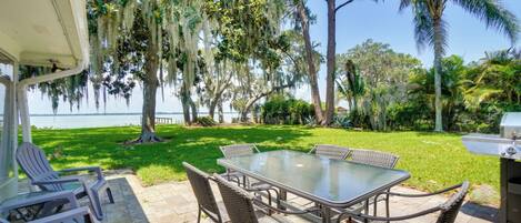 An outdoor patio with a glass table and wicker chairs, overlooking a grassy area and a body of water.