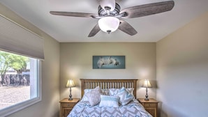 Un dormitorio con una cama, dos mesitas de noche con lámparas, un ventilador de techo, una ventana con persianas y un cuadro colgado en la pared.