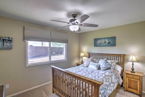 Une chambre avec un lit en bois, un ventilateur de plafond, une fenêtre avec des stores, une table de chevet avec une lampe et un tableau accroché au mur.