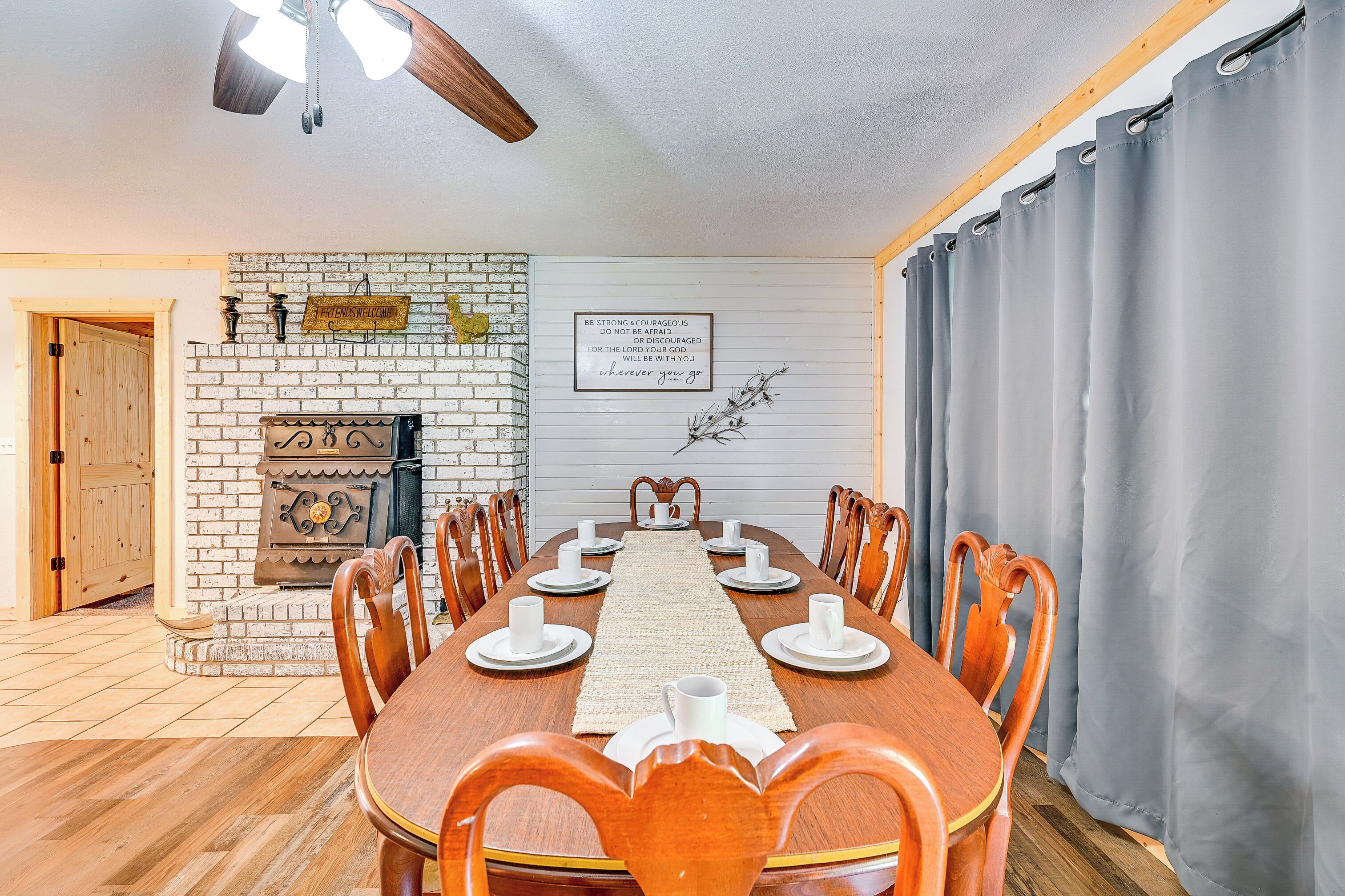 A dining room with a wooden table set for six, flanked by chairs, a brick fireplace, and a wall-mounted sign.