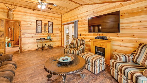 A cozy log cabin interior with a fireplace, striped armchairs, a round coffee table, and a mounted deer head.