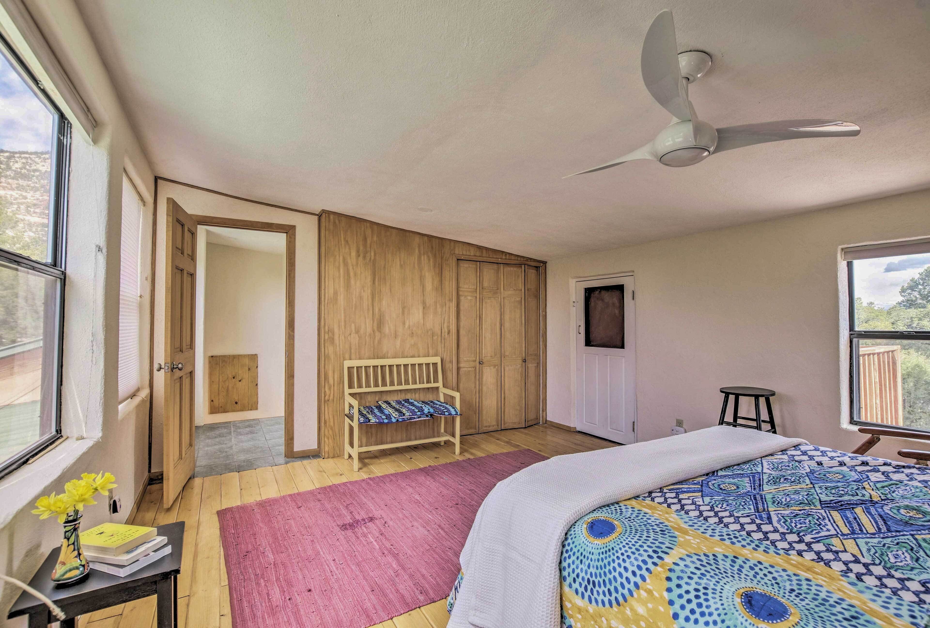 A bedroom with a bed, a ceiling fan, a wooden bench, a small stool, and a window with a view of the outdoors.