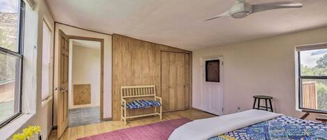 A bedroom with a bed, a ceiling fan, a wooden bench, a small stool, and a window with a view of the outdoors.