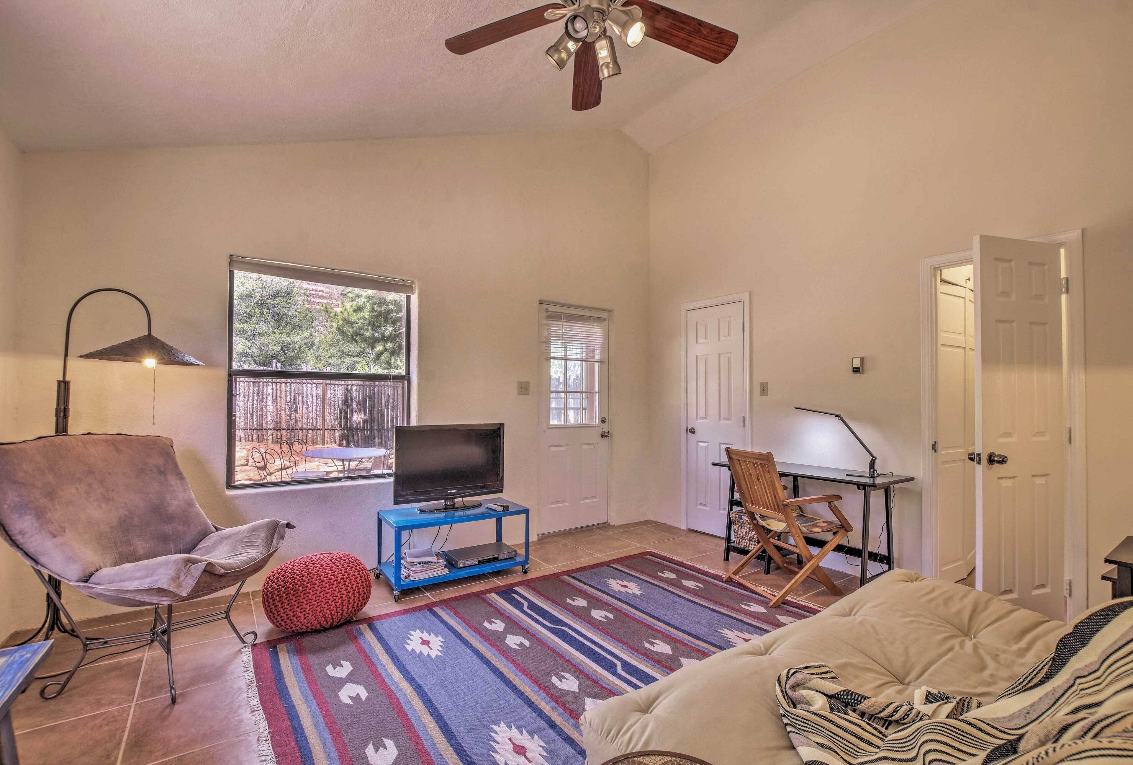 A living room with a ceiling fan, a TV on a blue stand, a red ottoman, a patterned rug, and a beige sofa.