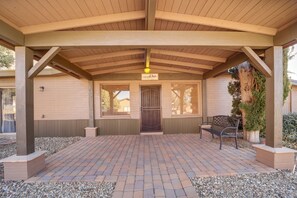 Un espacio al aire libre con techo, suelo de ladrillo, una pérgola de madera, un banco y una puerta con un letrero encima.